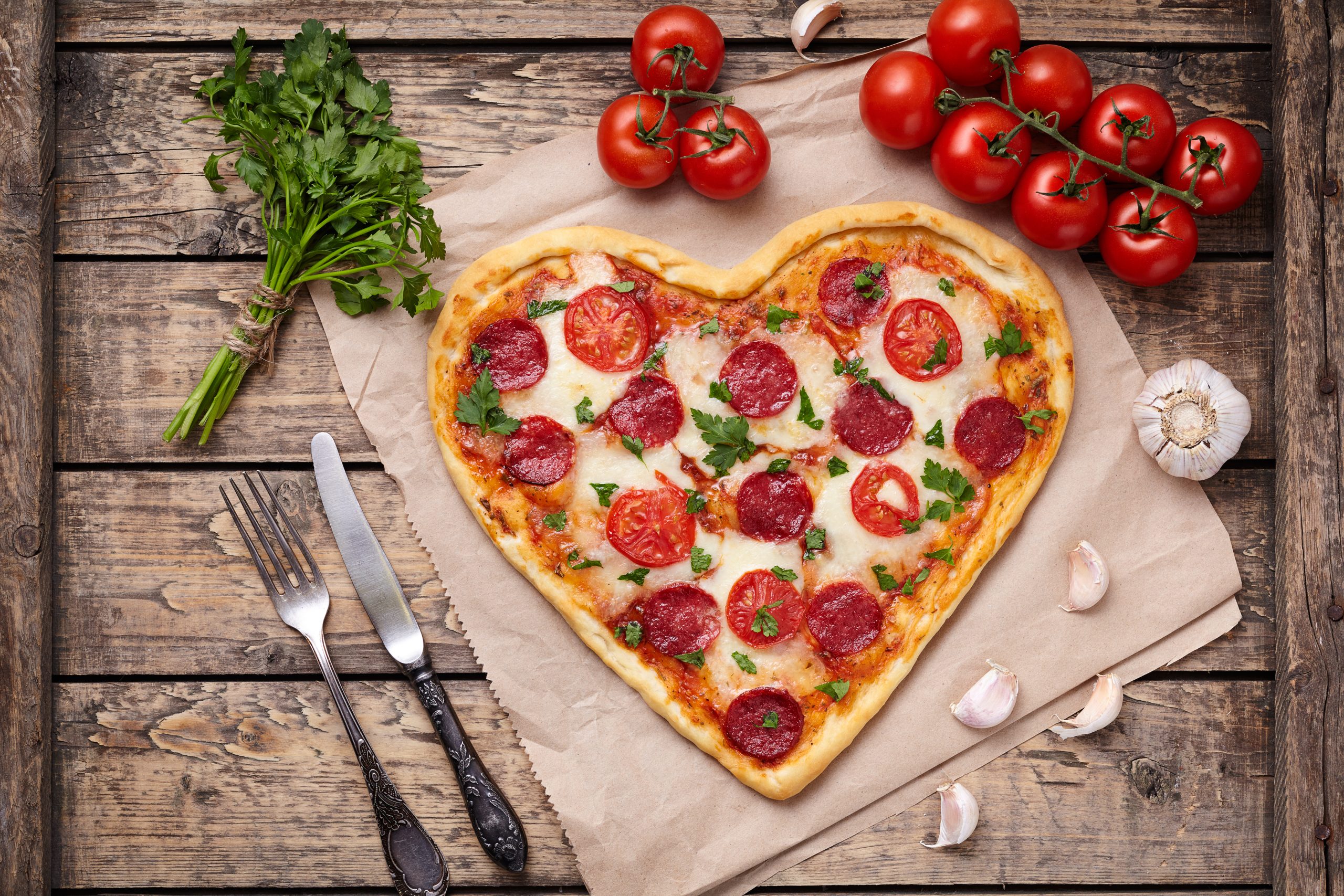 Heart shaped pizza for Valentines day with pepperoni, mozzarella, tomatoes, parsley and garlic on vintage wooden table background. Food symbol of romantic love.